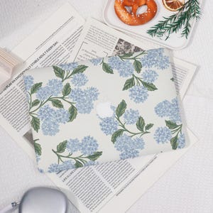 May include: A laptop case featuring a floral design with light blue hydrangea blooms and green leaves on a cream background. The case has a white Apple logo in the center. The case is on top of newspapers.
