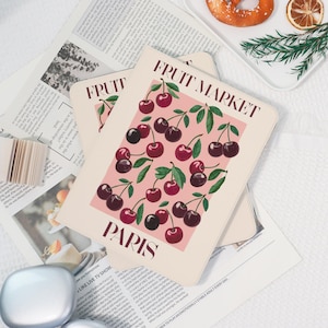 May include: A stack of three rectangular art prints with a pink background and a cherry illustration. The prints have the text "FRUIT MARKET" and "PARIS" in a dark red font. The prints are on top of a newspaper.