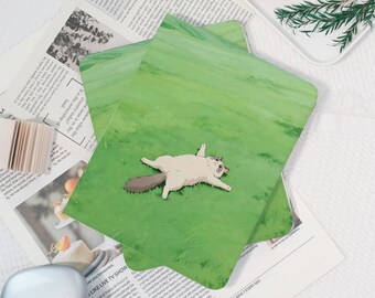 May include: Two green notebooks with a cartoon cat illustration on the cover, lying on a grassy field. The cat is light brown with a dark tail. The notebooks are on top of a newspaper.