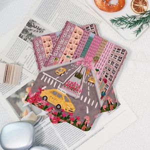 May include: A stack of New York City-themed art prints featuring yellow taxis, buildings, and floral accents. The prints are on a newspaper background, with a plate of food in the background.