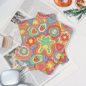 Könnte beinhalten: Ein sternförmiger Untersetzer mit einem bunten Retro-Design. Der Untersetzer zeigt einen grünen Frosch, lächelnde Blumen, Herzen und Sterne in einem lebendigen, wellenförmigen Muster. Der Untersetzer liegt auf einer Zeitung.