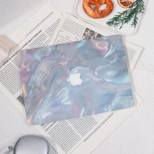 May include: A laptop with a pearlescent, iridescent cover featuring a swirling, liquid-like design in shades of blue, pink, and purple. The Apple logo is visible. The laptop rests on a background of newspaper clippings.