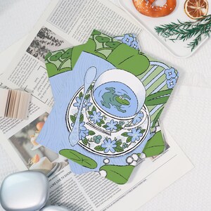 Könnte beinhalten: Eine skurrile Illustration einer Teetasse und Untertasse mit einem Frosch im Tee. Die Tasse und Untertasse sind mit blauen Blumenmustern verziert. Ein Löffel liegt auf der Untertasse. Der Hintergrund zeigt grüne Blätter und blaue Streifen.