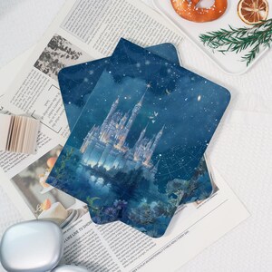 May include: Two blue greeting cards depicting a fairytale castle under a starry night sky. The cards are layered on a newspaper with text and images. A small stack of paper and a silver object are in the foreground.