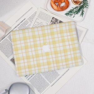 May include: A yellow and white plaid laptop case with the Apple logo in the center. The case is on top of newspaper clippings. A pretzel and a slice of dried orange are on a white plate in the background.