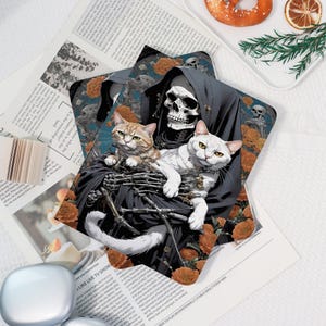 May include: A decorative print depicting a skeletal figure in a black cloak cradling two cats. One cat is ginger and white, the other is white. The background features orange flowers and skulls. The print is on a white surface with newspaper clippings.
