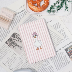Pode incluir: Caderno com capa listrada rosa com um ganso de desenho animado usando óculos de sol e uma faixa de cabelo floral. O caderno está em um fundo de recortes de jornal e um pretzel.