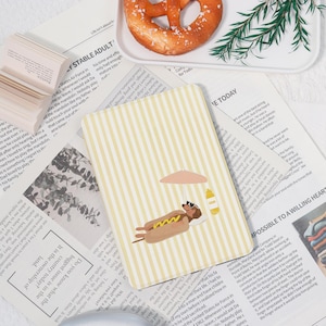 Peut inclure: Un bloc-notes rectangulaire à rayures verticales jaunes et blanches. Le bloc-notes présente une illustration d'un hot-dog se relaxant sous un parasol. Un bretzel et de la verdure sont sur un plateau blanc.