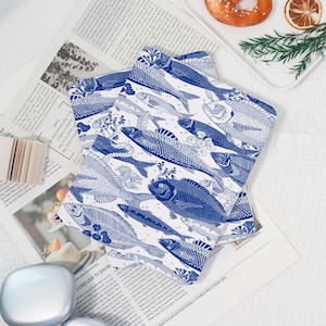 May include: Two square, white and blue patterned decorative cloths. The cloths feature a repeating design of various fish illustrations. The fish are in shades of blue, with detailed scales and fins, set against a white background. The cloths are placed on a newspaper.