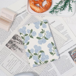 May include: A floral-patterned notepad featuring light blue hydrangea blooms and green leaves on a white background. The notepad is placed on top of a newspaper with text. A pretzel is on a white plate.