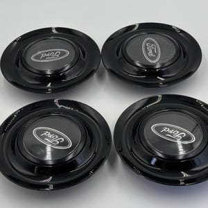 Könnte beinhalten: Vier schwarze Ford-Radkappen mit glänzendem Finish. Jede Radkappe hat das Ford-Logo in Weiß, zentriert auf einem erhabenen, kreisförmigen Bereich. Die Radkappen sind rund und scheinen neu zu sein.