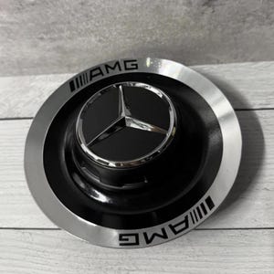 Könnte beinhalten: Eine schwarze und silberne AMG-Radnabenabdeckung mit dem Mercedes-Benz-Logo. Die Kappe hat eine schwarze Mitte mit einem silbernen Stern-Emblem und dem AMG-Logo in silberner Schrift.