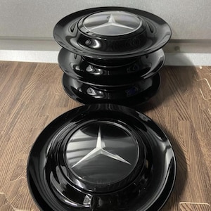 Könnte beinhalten: Vier schwarze Radkappen mit einem silbernen Mercedes-Benz-Logo, übereinander gestapelt. Eine Radkappe ist separat davor platziert. Die Radkappen haben eine glänzende Oberfläche und reflektieren das Licht.