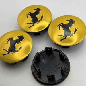 May include: Four gold-colored wheel center caps with a black prancing horse logo. One black plastic wheel cap base is also visible. The caps have a shiny finish and are likely for a car.