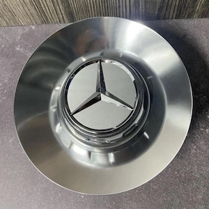 Könnte beinhalten: Eine glänzende, silberne Radkappe mit einem prominenten Mercedes-Benz-Logo in der Mitte. Das Logo zeigt einen dreizackigen Stern in einem kreisförmigen Emblem. Die Radkappe hat eine reflektierende, metallische Oberfläche und eine runde Form.