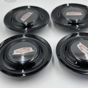 Könnte beinhalten: Vier schwarze Radkappen mit glänzendem Finish. Jede Radkappe hat eine transparente Mitte mit einem Cadillac-Emblem in Silber, Rot, Gold und Blau. Die Radkappen sind quadratisch auf weißem Hintergrund angeordnet.