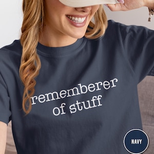 Puede incluir: Camiseta azul marino con el texto blanco "rememberer of stuff". La camiseta tiene cuello redondo y mangas cortas. Un círculo azul marino con la palabra "NAVY" está en la esquina inferior derecha.