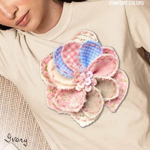 Peut inclure: T-shirt couleur ivoire orné d'une grande fleur faite à la main. La fleur présente des pétales en tissus roses, bleus et beiges, avec des motifs vichy et floraux. Le texte "COMFORT COLORS" est en haut et "Ivory" est écrit en bas.