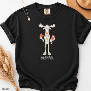 May include: A black short-sleeve t-shirt featuring a graphic of a white moose holding a muffin and a calculator. The moose wears a lanyard and the text below reads: Just One Muffin Was Never an Option. The shirt has a Comfort Colors brand label.