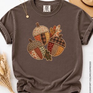 May include: A dark brown short-sleeve t-shirt featuring a central graphic of three acorns composed of various patterned fabric patches in shades of orange, brown, and cream, accented with two small leaves and a Comfort Colors brand label.