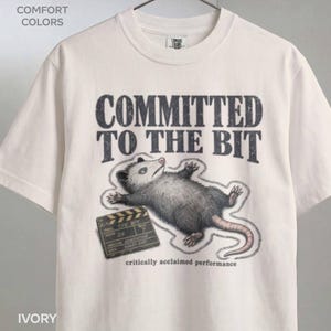 Peut inclure: Un t-shirt à manches courtes ivoire avec un motif représentant un opossum faisant le mort à côté d'un clap de cinéma. Le texte indique COMMITTED TO THE BIT et critically acclaimed performance. Le t-shirt porte une étiquette Comfort Colors.