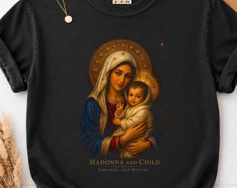 May include: A black short-sleeve t-shirt featuring a graphic print of the Madonna and Child with gold halos. Below the image, the text reads MADONNA AND CHILD and EMMANUEL, GOD WITH US. The shirt has a Comfort Colors brand label at the neck.