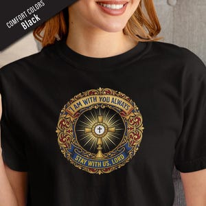 May include: Black t-shirt featuring a gold, red, and blue religious emblem. The emblem includes a cross, the text "I AM WITH YOU ALWAYS", and "STAY WITH US, LORD". The shirt is made of a comfortable material.