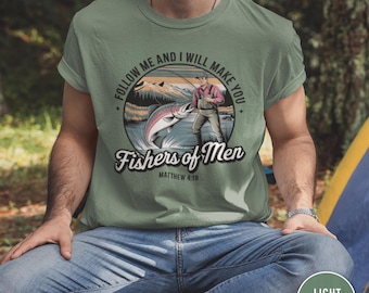Può includere: Una t-shirt verde chiaro con una grafica circolare che mostra una persona che pesca in un lago con montagne sullo sfondo. Il testo recita "FOLLOW ME AND I WILL MAKE YOU Fishers of Men MATTHEW 4:19" in un mix di caratteri diversi.