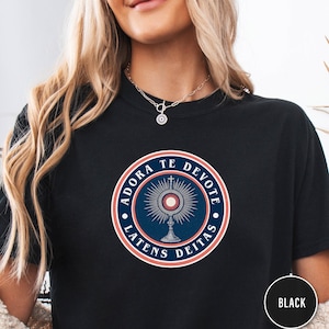 Pode incluir: Camiseta preta com um design circular. O design apresenta a frase latina "ADORA TE DEVOTE LATENS DEITAS" em torno de um símbolo religioso em azul, vermelho e branco. Um círculo preto com "BLACK" está no canto inferior direito.