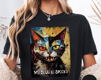 May include: Black t-shirt featuring a colorful, artistic cat face with one orange and one yellow eye. The text "MY SOUL IS SPOOKY" is below the cat. A black circle with the word "BLACK" is in the lower right corner.