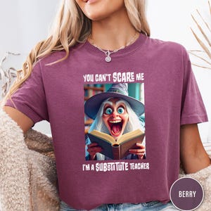 May include: Berry-colored t-shirt with the text "YOU CAN'T SCARE ME I'M A SUBSTITUTE TEACHER." The shirt features a graphic of a cartoon witch reading a book with a surprised expression. The shirt has short sleeves.