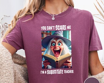 May include: Berry-colored t-shirt with the text "YOU CAN'T SCARE ME I'M A SUBSTITUTE TEACHER." The shirt features a graphic of a cartoon witch reading a book with a surprised expression. The shirt has short sleeves.