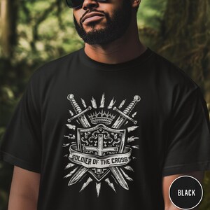 May include: Black t-shirt featuring a white graphic design. The design includes a shield, two crossed swords, a crown, and the text "SOLDIER OF THE CROSS" on a banner. The shirt is made of a soft material.
