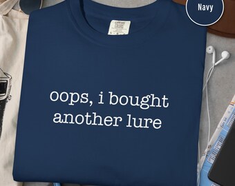 May include: Navy blue t-shirt with the text "oops, i bought another lure" in white. The shirt is a Comfort Colors brand. Sunglasses, a hat, and other items are in the background.