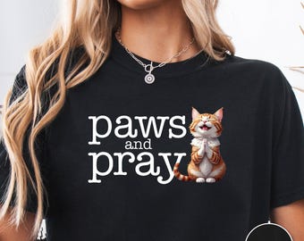 May include: Black t-shirt with the words "paws and pray" in white, accompanied by an illustration of a smiling orange tabby cat with its paws together. The shirt also has a black circle with the word "BLACK" in white.