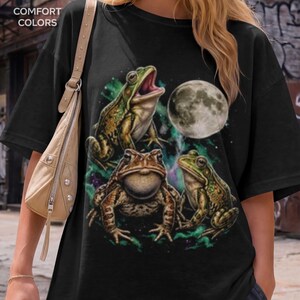 May include: A black t-shirt featuring a graphic print of three frogs and a full moon. The top frog has its mouth open, the middle frog has a puffed throat, and the third frog sits to the right. The text COMFORT COLORS is printed in the top left corner.