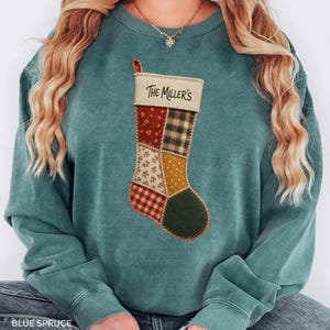 May include: A blue-green crewneck sweatshirt featuring a central graphic of a patchwork Christmas stocking. The stocking has a cream-coloured cuff with the text "THE MILLER'S" printed in black. The stocking body consists of various fabric patterns.
