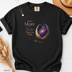 Könnte beinhalten: Ein schwarzes Kurzarm-T-Shirt mit einer Grafik eines aufgebrochenen Eies, das eine wirbelnde violette und orangefarbene Galaxie im Inneren zeigt. Der Text, There's More in You Than You Think, steht in hellbeiger Schrift neben dem Ei.