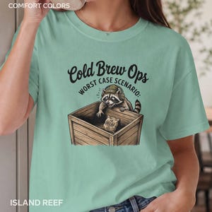 May include: A light green t-shirt featuring a graphic of a raccoon wearing headphones and a beanie, leaning into a wooden crate. The text reads Cold Brew Ops Worst Case Scenario and Decaf on a small bag inside the crate. The shirt is labeled Comfort Colors.
