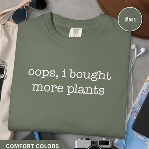 Pode incluir: Uma camiseta verde musgo com o texto branco "oops, I bought more plants". A camisa é da Comfort Colors. Óculos de sol, um chapéu e uma câmera são visíveis no fundo.