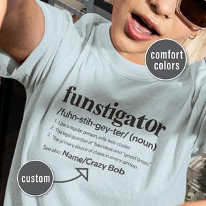 May include: Pale blue t-shirt with the word "funstigator" in black. The shirt has a definition of the word, including pronunciation and meaning. The text "comfort colors" and "custom" are also visible on the shirt.