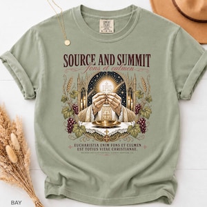 May include: A sage green crew neck t-shirt featuring a graphic print of hands holding a communion host above a chalice. The text reads "SOURCE AND SUMMIT" and "EUCHARISTIA ENIM FONS ET CULMEN EST TOTIUS VITAE CHRISTIANAE" with ornate gothic arch details.