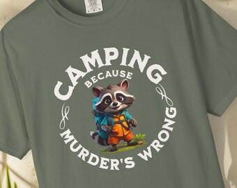 May include: A moss-colored t-shirt with a graphic of a raccoon wearing a backpack. The text reads "CAMPING BECAUSE MURDER'S WRONG" in a circular design around the raccoon. The shirt is Comfort Colors brand, unisex sizing.