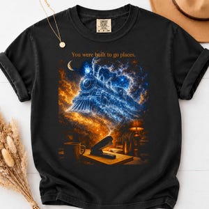 May include: A black t-shirt featuring a graphic print of a steam locomotive emerging from a nebula of blue and orange cosmic clouds. Above the train, the text reads, "You were built to go places." Below the train, a desk scene includes a mug and stapler.