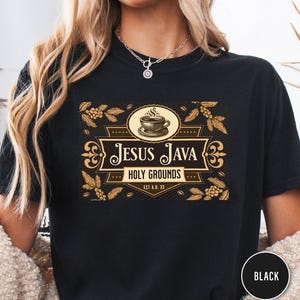 Op de afbeelding: Zwart t-shirt met een koffie-thema ontwerp. Het ontwerp bevat de tekst "Jesus Java Holy Grounds" met een illustratie van een koffiekopje en decoratieve elementen. Op het shirt staat ook "EST A.D. 33".