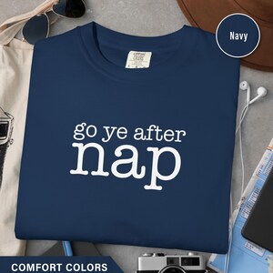 Könnte beinhalten: Marineblaues T-Shirt mit dem weißen Schriftzug "go ye after nap", einem Comfort Colors-Etikett und einem marinefarbenen Farbmuster. Das T-Shirt wird mit Accessoires wie Sonnenbrille, Hut und Kamera präsentiert.