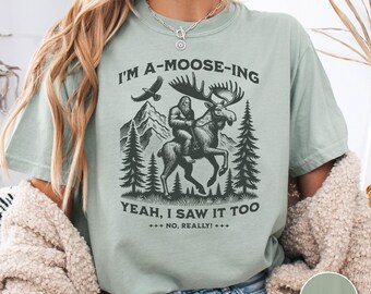 Może przedstawiać: Szałwiowo-zielony t-shirt z krótkim rękawem i ciemnozieloną grafiką przedstawiającą stworzenie jadące na łosiu w górskim lesie. Napis głosi I'M A-MOOSE-ING YEAH, I SAW IT TOO NO, REALLY! Koszulka ma okrągły dekolt i luźny krój.