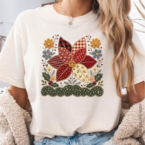 Könnte beinhalten: Ein elfenbeinfarbenes T-Shirt mit einem zentralen Motiv einer großen Weihnachtssternblüte, die aus verschiedenen roten, cremefarbenen und karierten stoffartigen Blütenblättern besteht. Das Design umfasst gelbe Blumen und grüne Blätter.