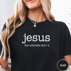 Può includere: T-shirt nera con la parola "jesus" in bianco, seguita da "the ultimate ctrl + z". La maglietta ha un girocollo e maniche corte. Un cerchio nero con la scritta "BLACK" si trova in basso a destra.