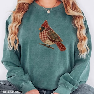 May include: A blue-green crewneck sweatshirt featuring a central graphic of a cardinal bird. The bird is composed of various fabric-like patches in red, brown, and cream with plaid and floral patterns, perched on a small brown branch.